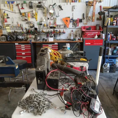 Assorted workshop tools and equipment, including a heavy-duty chain, battery chargers, and hand tools, displayed on a folding table in a...
