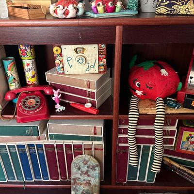 Sale Photo Thumbnail #66: Collection of vintage toys and books on wooden shelves, including a red rotary toy telephone, a Fisher-Price music box, a plush tomato character with striped legs, and various classic hardback books.