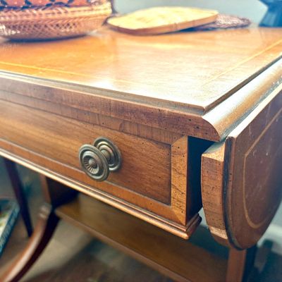 Sale Photo Thumbnail #94: Vintage drop-leaf wooden side table featuring a single drawer with a decorative brass pull and inlaid border detailing.