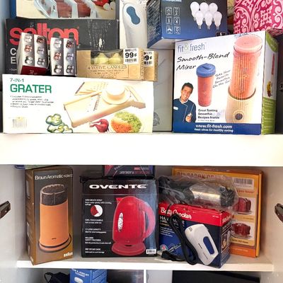 Sale Photo Thumbnail #93: Assorted small kitchen appliances and household items in their original boxes, including a Braun coffee grinder, Ovente electric kettle, and Hamilton Beach hand mixer.