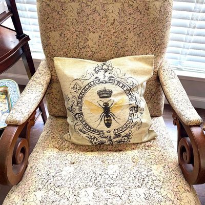 Sale Photo Thumbnail #100: Vintage upholstered armchair featuring a pink and cream floral pattern, wooden scroll arms, and a decorative throw pillow with a crowned bee motif.