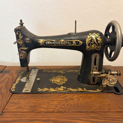 Sale Photo Thumbnail #70: Antique Minnesota Model A treadle sewing machine head with ornate gold filigree decals, mounted in a wooden cabinet.