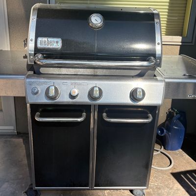 Weber Genesis three-burner gas grill with a black porcelain-enameled lid, stainless steel side tables, and a built-in thermometer.