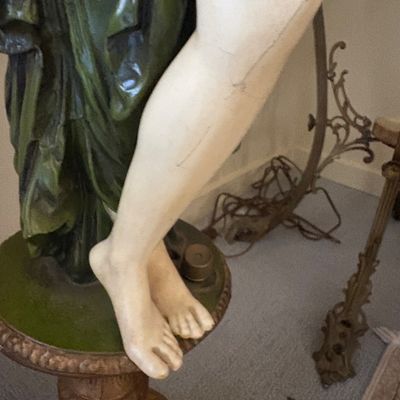 Detail view of a classical-style female statue showing a hairline crack on the leg, standing on an ornate pedestal.