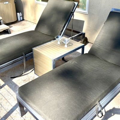 Pair of modern outdoor chaise lounges with dark grey cushions, paired with a matching slatted side table.
