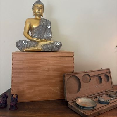 Decorative Buddha statue with gold and silver-toned finish sitting atop a wooden box, displayed alongside an antique wooden scale set in...