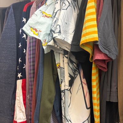 Assorted men's clothing hanging in a closet, including graphic tees, button-down shirts, and a long-sleeve shirt with an American...