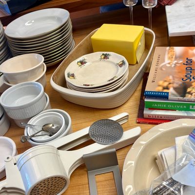Assorted kitchenware including stacks of white plates, small ramekins, a white potato ricer, cookbooks, and patterned dessert plates.