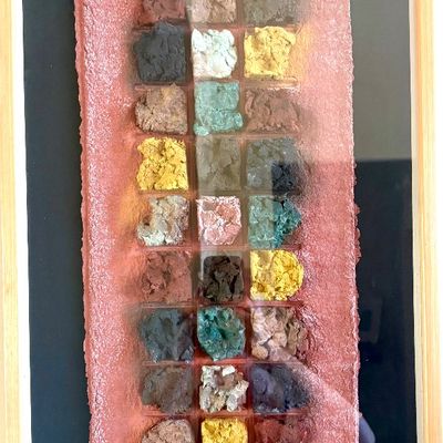Framed mixed-media textile art piece featuring a grid of textured, colorful earth-toned squares on a pinkish-red background, presented in...