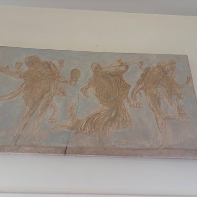 Classical-style plaster wall relief plaque depicting three dancing figures, showing some cracking and wear.