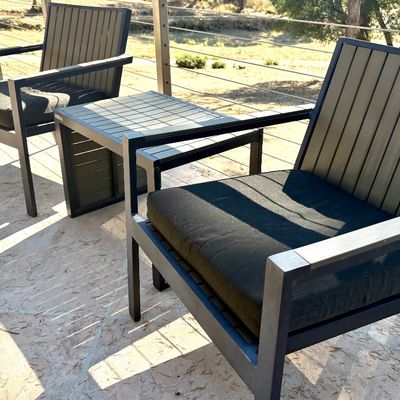 Modern outdoor patio set featuring two dark metal armchairs with slatted backs, black seat cushions, and a matching small side table.