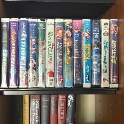 Collection of vintage VHS tapes, including Disney classics and family movies, displayed on wooden shelves alongside various hardcover books.