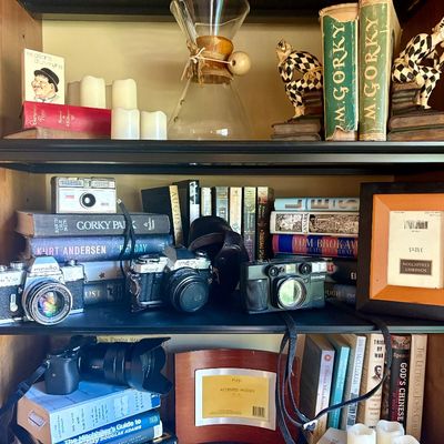 A collection of vintage cameras, books, and decorative items displayed on wooden shelves, including a Minolta SRT 101 SLR camera and a...