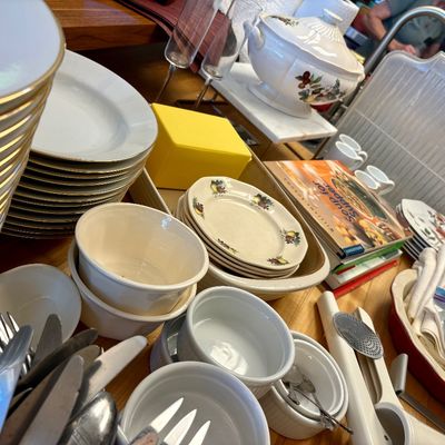 Assorted kitchenware and dining items, including gold-rimmed plates, ceramic bowls, a soup tureen, cookbooks, and flatware.