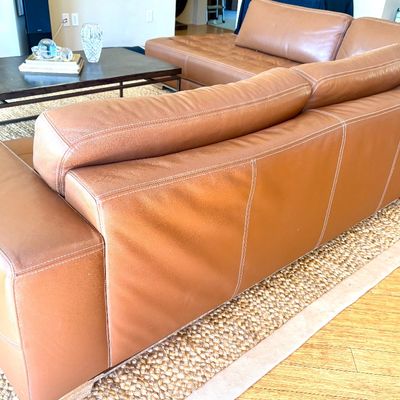 Contemporary tan leather sectional sofa with white contrast stitching, shown from the back, paired with a low-profile coffee table in a...