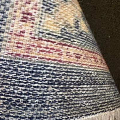 Close-up view of the underside of a woven area rug, showing the tight knotting, fringe detail, and a pattern in shades of blue, red, and...