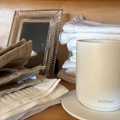 Ember smart mug in white with its charging coaster, displayed alongside folded linens and a silver-toned picture frame.
