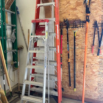 Werner aluminum and fiberglass A-frame ladders shown alongside various long-handled yard tools, including a Fiskars pole saw and loppers.
