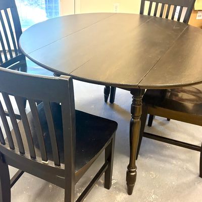 Black wooden drop-leaf dining table with turned legs, accompanied by four matching mission-style slat-back chairs.