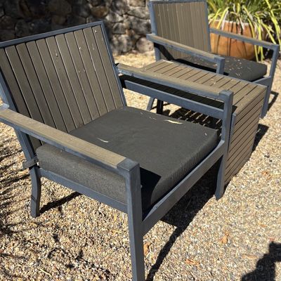 Modern outdoor patio set featuring two armchairs with dark grey metal frames, brown composite wood slats, and black seat cushions, paired...