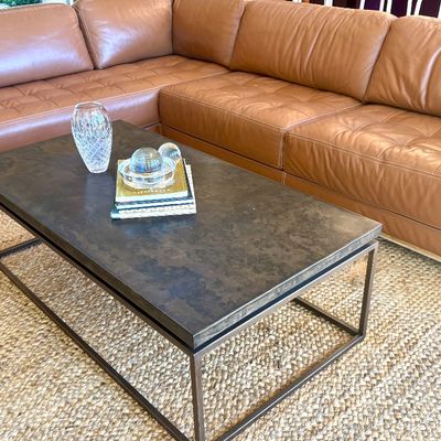 Modern rectangular coffee table with a dark stone-look top and a dark metal frame, shown in front of a tan leather sectional sofa.