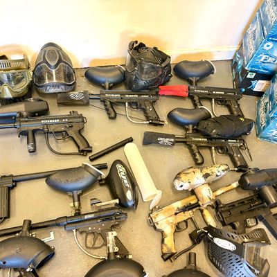 Collection of paintball gear, including Tippmann markers, hoppers, protective masks, and cases of Triumph paintballs.