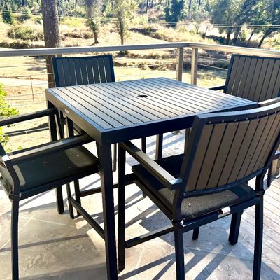 Modern outdoor dining set featuring a square slatted table with an umbrella hole and four matching armchairs with black seat cushions.