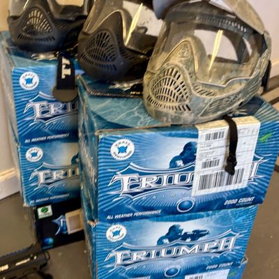 Three paintball masks in black and camouflage patterns, displayed on top of stacked boxes of Triumph paintballs.