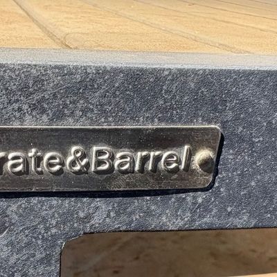 Crate & Barrel metal logo plaque affixed to the dark grey frame of an outdoor table with a light wood-look slatted top.