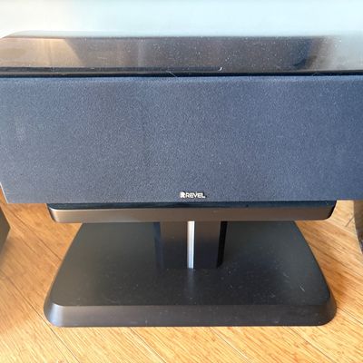 Revel center channel speaker with a black fabric grille, mounted on a matching black pedestal stand.