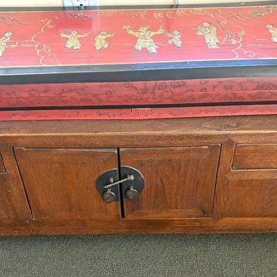 Asian-style wooden low cabinet with metal hardware, topped with a red and black lacquered box featuring hand-painted figures.