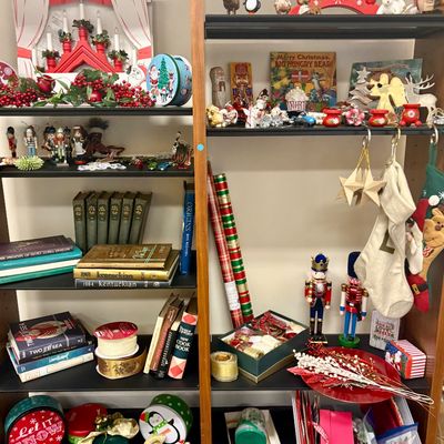 A collection of Christmas decorations, books, and wrapping supplies displayed on a wooden shelving unit.