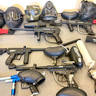 Collection of paintball gear, including Tippmann markers, hoppers, and protective face masks in various colors and styles.