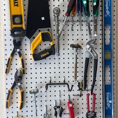 Assorted hand tools organized on a white pegboard, including DeWalt and Empire levels, a hand saw, screwdrivers, pliers, hammers, and...