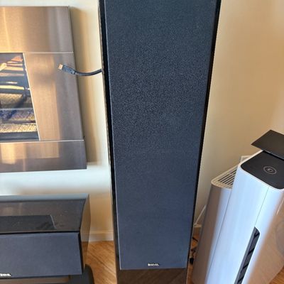 Revel floor-standing tower speaker with a glossy black finish and a black fabric grille, shown alongside other home audio equipment.