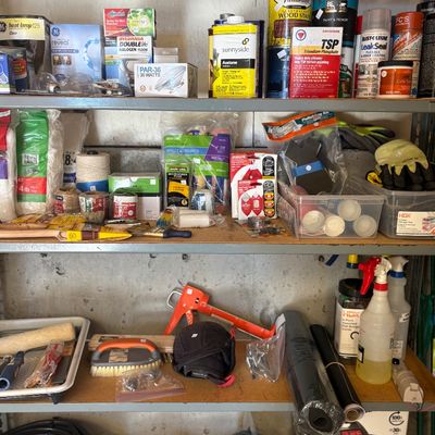 Assorted garage and workshop supplies on metal shelving, including paint, solvents, lightbulbs, hand tools, and cleaning products.