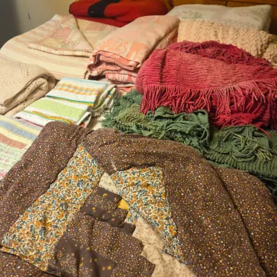 Sale Photo Thumbnail #45: Assortment of vintage blankets, quilts, and throws, including a brown floral patchwork quilt, fringed knit blankets, and striped towels arranged on a wooden bed.