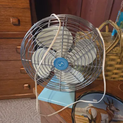Sale Photo Thumbnail #7: Vintage mid-century electric desk fan featuring a light blue base, four metal blades, and a chrome wire safety cage with a blue center emblem.