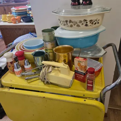 Sale Photo Thumbnail #54: Vintage yellow metal rolling utility cart loaded with mid-century kitchenware, including Pyrex mixing bowls, a hand mixer, anodized aluminum cups, and novelty salt and pepper shakers.