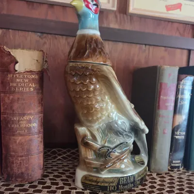 Sale Photo Thumbnail #95: Vintage Jim Beam ceramic pheasant decanter, 110 months old Kentucky Straight Bourbon Whiskey, displayed on a shelf alongside antique books.
