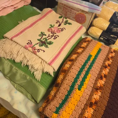 Sale Photo Thumbnail #51: Assortment of vintage blankets and craft supplies, featuring a hand-crocheted afghan in earth tones, a green thermal blanket with satin trim, and a fringed throw with embroidered floral details.
