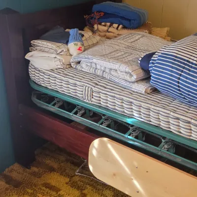 Sale Photo Thumbnail #34: Vintage wooden single bed frame with an exposed green metal spring base, topped with a striped mattress and various folded blankets.