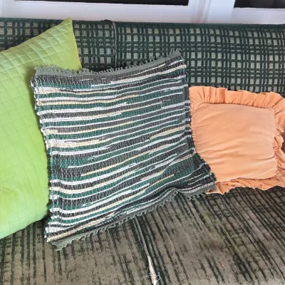 Sale Photo Thumbnail #17: Assorted throw pillows, including a green quilted square pillow, a woven rag-style striped pillow, and a peach velvet ruffled pillow, displayed on a green patterned sofa.