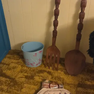Sale Photo Thumbnail #30: Vintage giant carved wooden fork and spoon wall decor set with tiki-style handles, shown alongside a light blue floral wastebasket and a woven handbag.