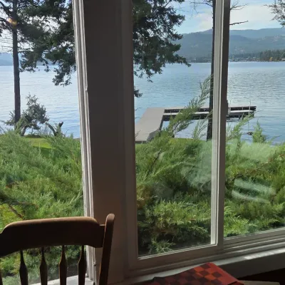 Sale Photo Thumbnail #23: Scenic lake view through a large window, featuring a wooden dock, evergreen trees, and a partial view of a wooden dining chair in the foreground.