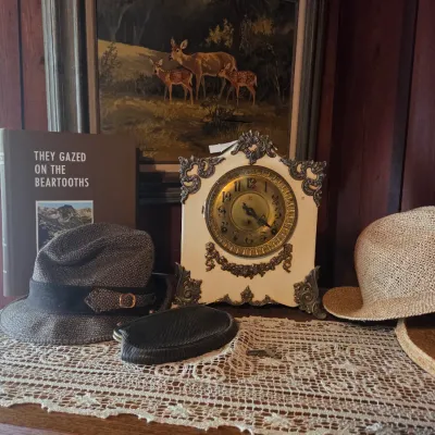 Sale Photo Thumbnail #79: Vintage mantel clock with ornate metal filigree, displayed alongside a copy of "They Gazed on the Beartooths," a tweed fedora, a straw hat, and a framed deer painting.