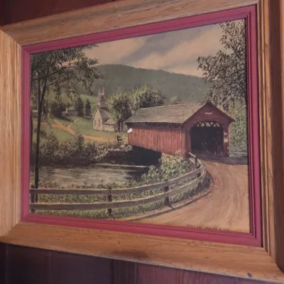 Sale Photo Thumbnail #76: Framed landscape painting depicting a red covered bridge and a church in a rural setting, housed in a wooden frame with a red inner border.
