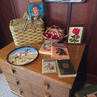 Sale Photo Thumbnail #82: Vintage collection of items on a wooden dresser, including a metal picnic basket, a decorative tray featuring an antelope, binoculars in a leather case, a framed rose embroidery, and several vintage books.