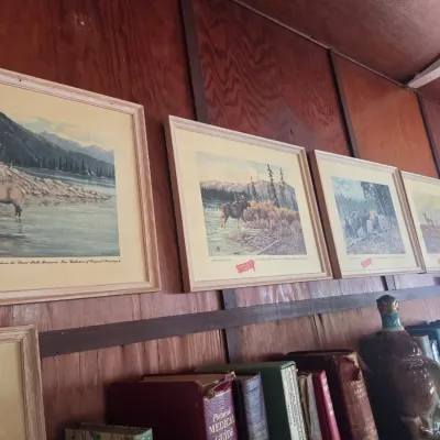 Sale Photo Thumbnail #96: Four framed vintage Great Falls Select beer advertising prints featuring wildlife scenes, mounted on a wood-paneled wall above a shelf of books.