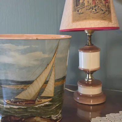 Sale Photo Thumbnail #40: Vintage metal wastebasket featuring a sailboat illustration, paired with a mid-century ceramic table lamp with a fox hunting scene on the shade.